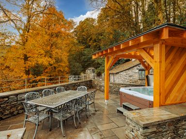 An outdoor patio with a metal dining table and chairs and a wooden covered hot tub surrounded by trees at Gate House in Coniston