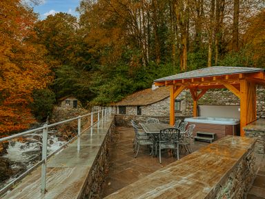 An outdoor patio area with metal chairs and tables next to a hot tub under a wooden gazebo by a stone wall and river at Gate House in Coniston