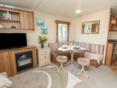 A dining area with a corner bench and stools around a table set with plates and glasses a television above a fireplace and a small drawer unit with flowers at 15 Rosewood in Great Thorness near Northwood Isle Of Wight