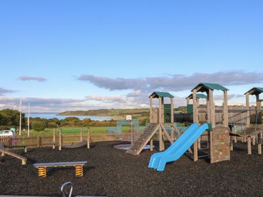 An outdoor playground with wooden climbing structures and blue slides near a fenced green field and water at 15 Rosewood Great Thorness near Northwood Isle Of Wight
