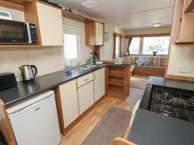 A kitchen area with a microwave, kettle, toaster, sink, stove, and fridge leading into a lounge with a corner sofa and windows at 15 Rosewood Great Thorness near Northwood Isle Of Wight