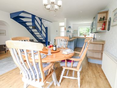 A kitchen and dining area with a wooden table set with plates and a bottle near a blue staircase at Mermaid Cottage in Wakeham Portland