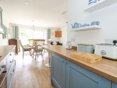 A kitchen with blue cabinets and wooden countertops next to a dining area with wooden chairs and curtains at Mermaid Cottage in Wakeham Portland