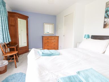 A bedroom with a bed covered with white sheets and blue towels a wooden wardrobe and chest of drawers and two wooden chairs with blue curtains at Mermaid Cottage in Wakeham Portland