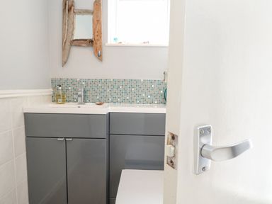 A bathroom with a grey cabinet sink and a mosaic tile backsplash a wooden framed mirror and a partially open white door at Mermaid Cottage in Wakeham Portland