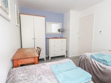 A bedroom with two beds covered with gray blankets and blue towels a wooden desk with a chair a white wardrobe and a white chest of drawers with a lamp and mirror on a blue accent wall at Mermaid Cottage in Wakeham Portland