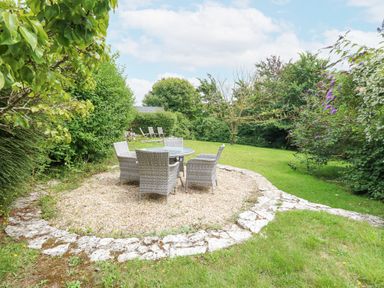 An outdoor seating area with a round glass table and four wicker chairs on gravel surrounded by grass and bushes at Mermaid Cottage in Wakeham Portland