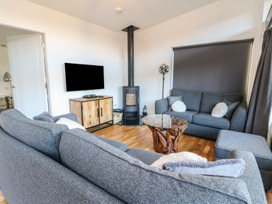 A living room with gray sofas a wooden cabinet a wall-mounted tv a wood-burning stove and a round glass coffee table at Osprey View Lodge & Hot Tub Aberfeldy