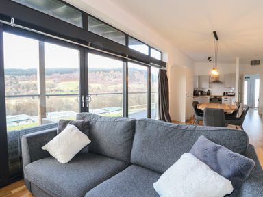 A living room with a gray sofa and cushions next to large windows showing a landscape at Osprey View Lodge & Hot Tub in Aberfeldy