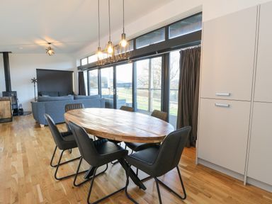 A dining area with a wooden table and six black chairs next to large windows and a living room with a gray sofa at Osprey View Lodge & Hot Tub in Aberfeldy