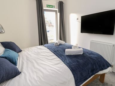 A bedroom with a bed covered with white and blue bedding and rolled towels on top a wall-mounted television and a fire exit door with curtains at Osprey View Lodge & Hot Tub Aberfeldy