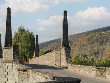 A stone bridge with four tall pillars over a road with trees and a forested hill in the background at Osprey View Lodge & Hot Tub in Aberfeldy