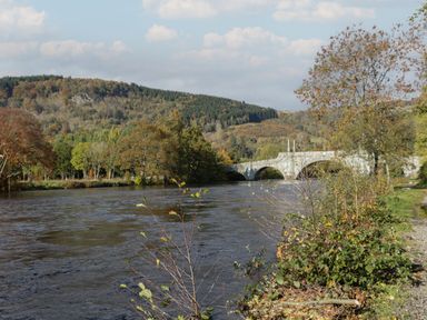 A river with a stone bridge surrounded by trees and hills at Osprey View Lodge & Hot Tub in Aberfeldy