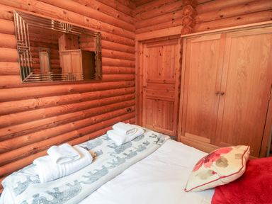 A bedroom with a bed towels a wooden wardrobe a door and a mirror on log cabin walls at 10 Waters Edge Morpeth near Felton