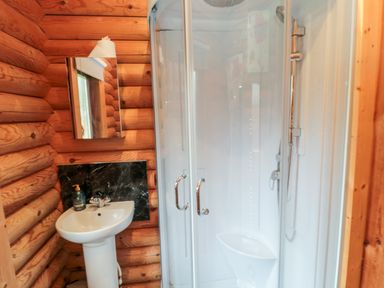 A bathroom with a white pedestal sink a mirror a soap dispenser and a curved glass shower enclosure with wooden log walls at 10 Water's Edge in Morpeth near Felton