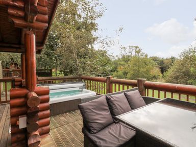 An outdoor deck with a hot tub a wooden railing and a table with cushioned chairs surrounded by trees at 10 Water's Edge Morpeth near Felton