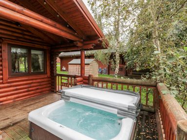 An outdoor hot tub on a wooden deck attached to a log cabin surrounded by trees at 10 Waters Edge in Morpeth near Felton