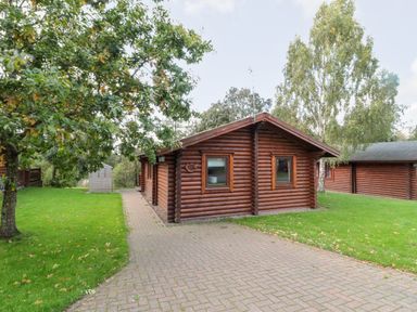 A wooden log cabin with a paved pathway and green lawn with trees at 10 Water's Edge in Morpeth near Felton