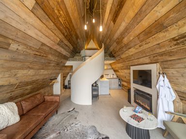 An interior view of an A-frame room with wooden walls a brown sofa a spiral staircase a kitchen a fireplace and a small table at The Tri-Pod Pantcefn near Llanarth