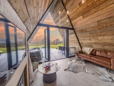 An interior living space with wooden walls a brown sofa a small round table and glass doors opening to an outdoor deck with a hot tub and chairs at The Tri-Pod in Pantcefn near Llanarth