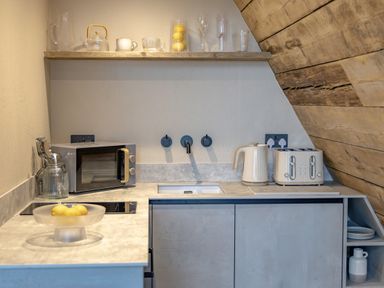 A small kitchen corner with a microwave kettle toaster sink and shelf with cups and glassware at The Tri-Pod in Pantcefn near Llanarth