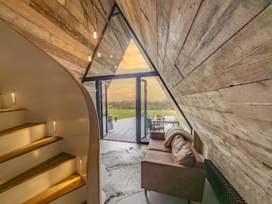 An interior space with wooden walls and stairs a brown sofa and glass doors opening to a deck and outdoor seating at The Tri-Pod in Pantcefn near Llanarth
