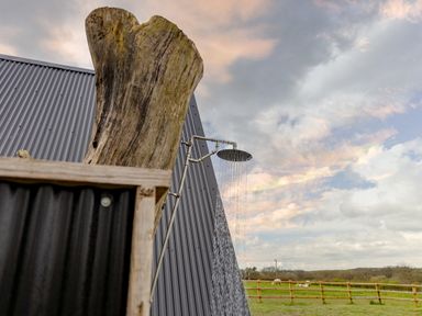 An outdoor shower with water running next to a wooden post and a metal building with a grassy field and wooden fence in the background at The Tri-Pod in Pantcefn near Llanarth