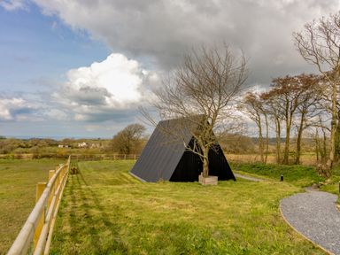 A black triangular cabin near a tree on a grassy field with a wooden fence and gravel path at The Tri-Pod in Pantcefn near Llanarth