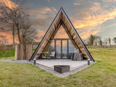 An outdoor view of an A-frame cabin with a table, chair, and hot tub on the porch at The Tri-Pod in Pantcefn near Llanarth