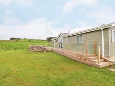 An exterior view of a single-story building with a wooden deck and glass railing on a grassy area at No.7 Skiddaw View in Moota near Aspatria