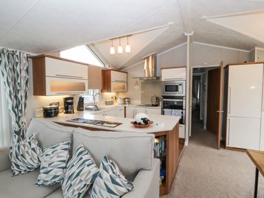 A kitchen with white cabinets and appliances adjacent to a living area with a grey sofa and patterned cushions at No.7 Skiddaw View in Moota near Aspatria