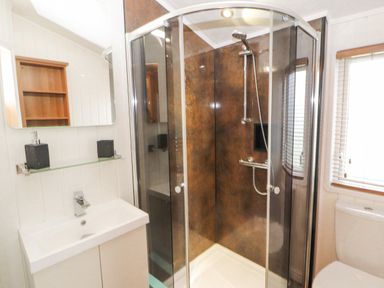 A bathroom with a glass shower enclosure a white sink with cabinet a mirror and a window with blinds at No 7 Skiddaw View Moota near Aspatria