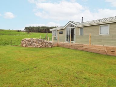 A single-story house with a stone wall and a wooden deck in a grassy area at No 7 Skiddaw View in Moota near Aspatria
