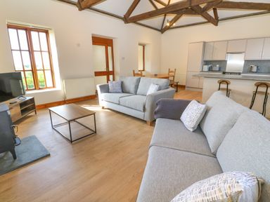 A living room with two gray sofas a coffee table a television a wood burner a dining table and a kitchen area with bar stools at The Granary in Prussia Cove near Rosudgeon