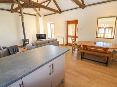An open plan living and dining area with wooden beams on the ceiling sofas a television wood stove dining table chairs and kitchen island at The Granary Prussia Cove near Rosudgeon