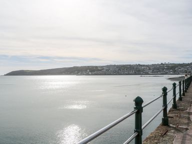 A metal railing along a stone path by a calm body of water with a distant shoreline and houses at The Granary Prussia Cove near Rosudgeon