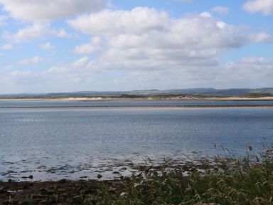A coastal landscape with water in the foreground and sand dunes in the background under a cloudy sky at Kits Cottage in Middleton near Belford