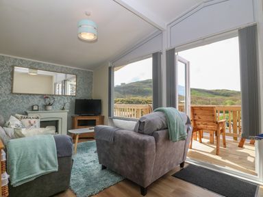 A living room with armchairs blankets a rug a TV a coffee table and a balcony with wooden chairs at Y Goedlan in Tywyn