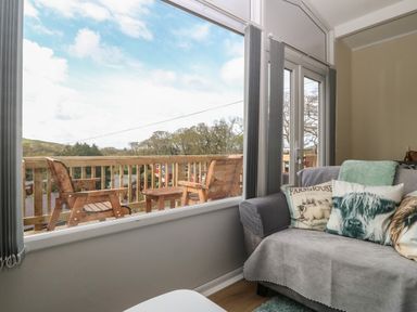 A living room with a sofa and decorative cushions next to a large window showing a balcony with wooden chairs at Y Goedlan in Tywyn