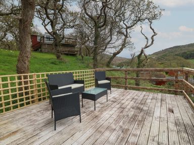 An outdoor wooden deck with outdoor furniture including chairs a table and a bench surrounded by trees at Y Goedlan in Tywyn