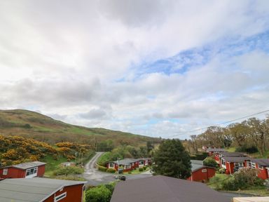 A cluster of red single-story houses with trees and hills in the background at Y Goedlan in Tywyn