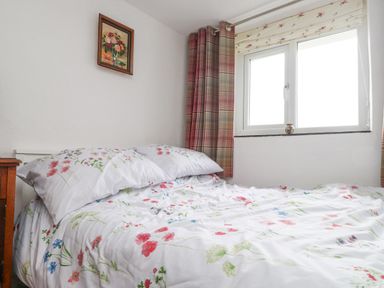 A bedroom with a bed having floral bedding beside a window with curtains and a framed painting on the wall at Quarry Cottage in Bethesda