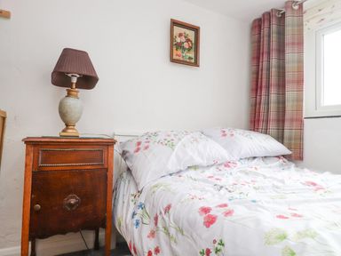 A bedroom with floral bed sheets a wooden bedside table with a lamp a small framed painting and a window with plaid curtains at Quarry Cottage in Bethesda