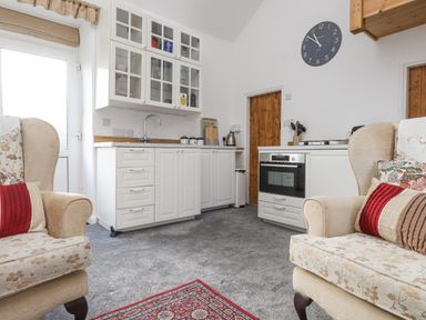 A kitchen area with white cabinets a sink an oven two armchairs and a red rug at Quarry Cottage in Bethesda
