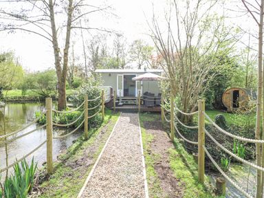 A gravel pathway with rope fencing leading to a small hut with outdoor seating and an umbrella next to a pond at Damsel Shepherds Hut in Badwell Ash