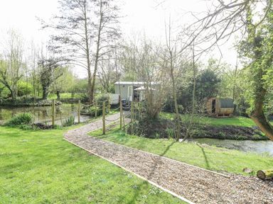 A gravel pathway leading to a small green hut and a wooden barrel sauna near a pond surrounded by trees at Damsel Shepherds Hut in Badwell Ash