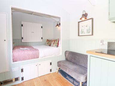 A built-in bed with colorful pillows and a pink blanket next to a gray velvet sofa in a small living area at Damsel Shepherds Hut in Badwell Ash