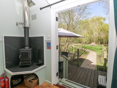 A wood-burning stove next to an open door leading to a garden path with an umbrella and trees at Damsel Shepherds Hut in Badwell Ash