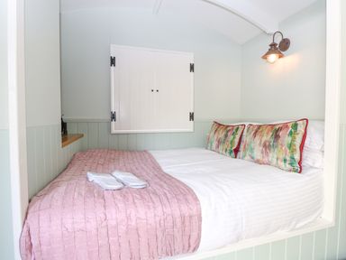 A small bedroom with a bed covered with white sheets and a pink blanket with colorful pillows and a wall light at Damsel Shepherds Hut in Badwell Ash