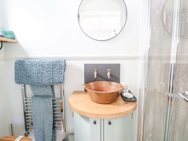 A bathroom with a round mirror above a wooden countertop basin and two taps a towel rack with blue towels and a glass shower door at Damsel Shepherds Hut in Badwell Ash